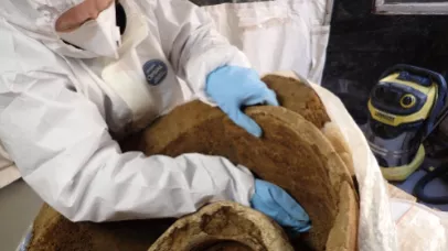 Fouille fine d'une urne en plomb. L'anthropologue porte une blouse intégrale, des gants et un masque pour se protéger.